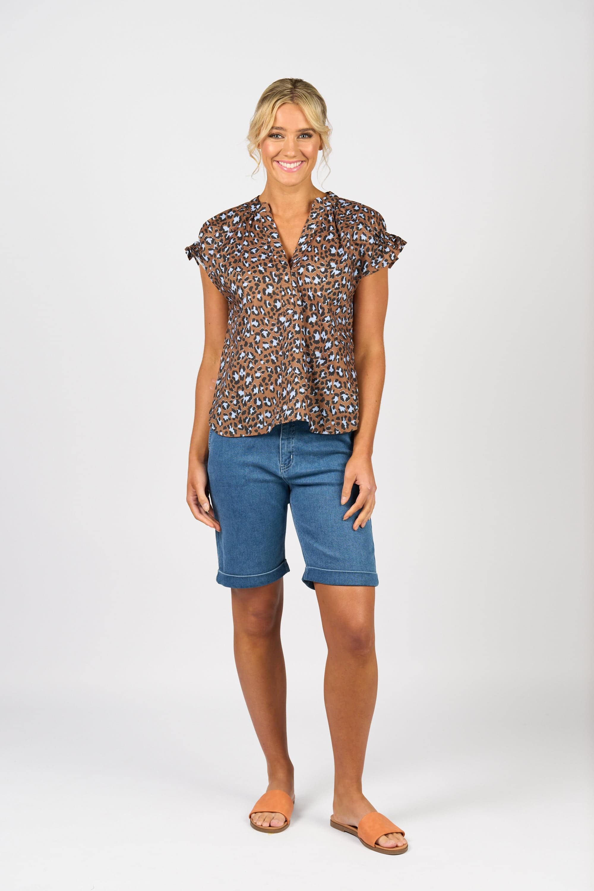 A woman stands smiling against a plain white background, wearing the Vassalli Evelyn Top - Nala with a brown and black leopard print, blue knee-length denim shorts, and tan sandals.
