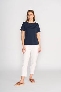 A woman with straight brown hair and a neutral expression wears the Vassalli Raya Top - Ink, a comfortable navy boat neck cotton-elastane top, paired with white pants and brown sandals against a plain light background.