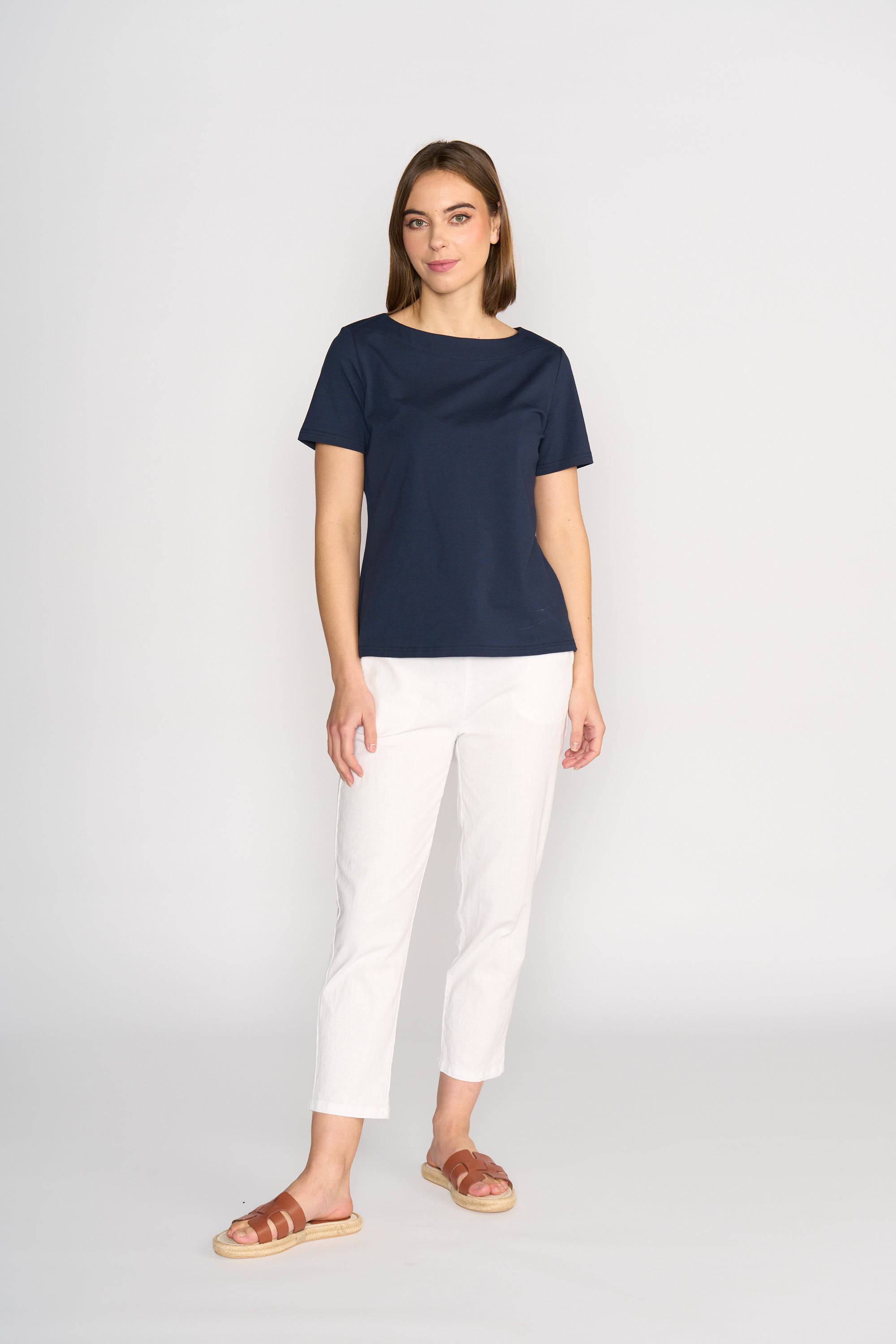 A woman with straight brown hair and a neutral expression wears the Vassalli Raya Top - Ink, a comfortable navy boat neck cotton-elastane top, paired with white pants and brown sandals against a plain light background.