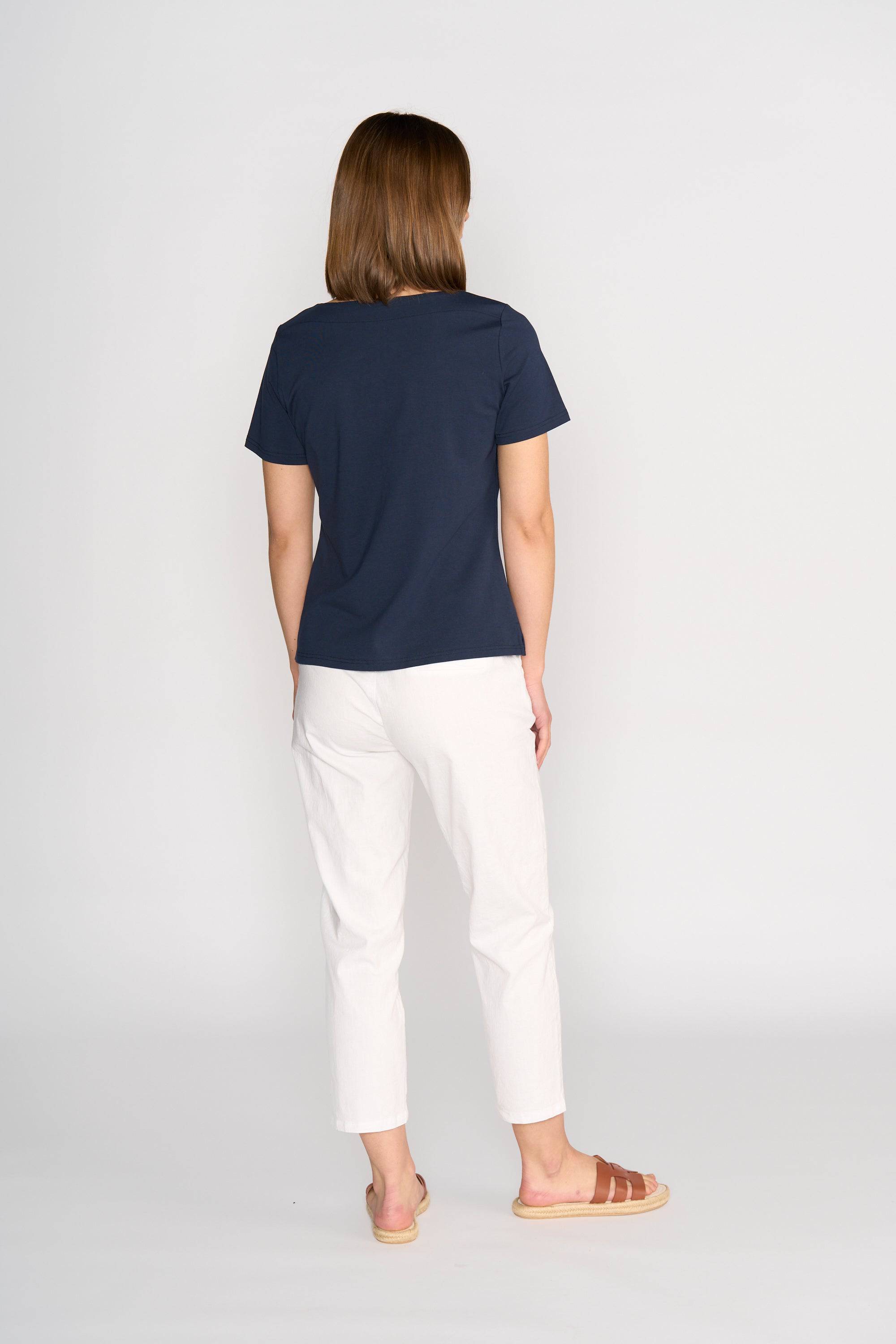 A woman faces away from the camera, wearing the Vassalli Raya Top - Ink, paired with white pants and brown sandals, against a plain light background.