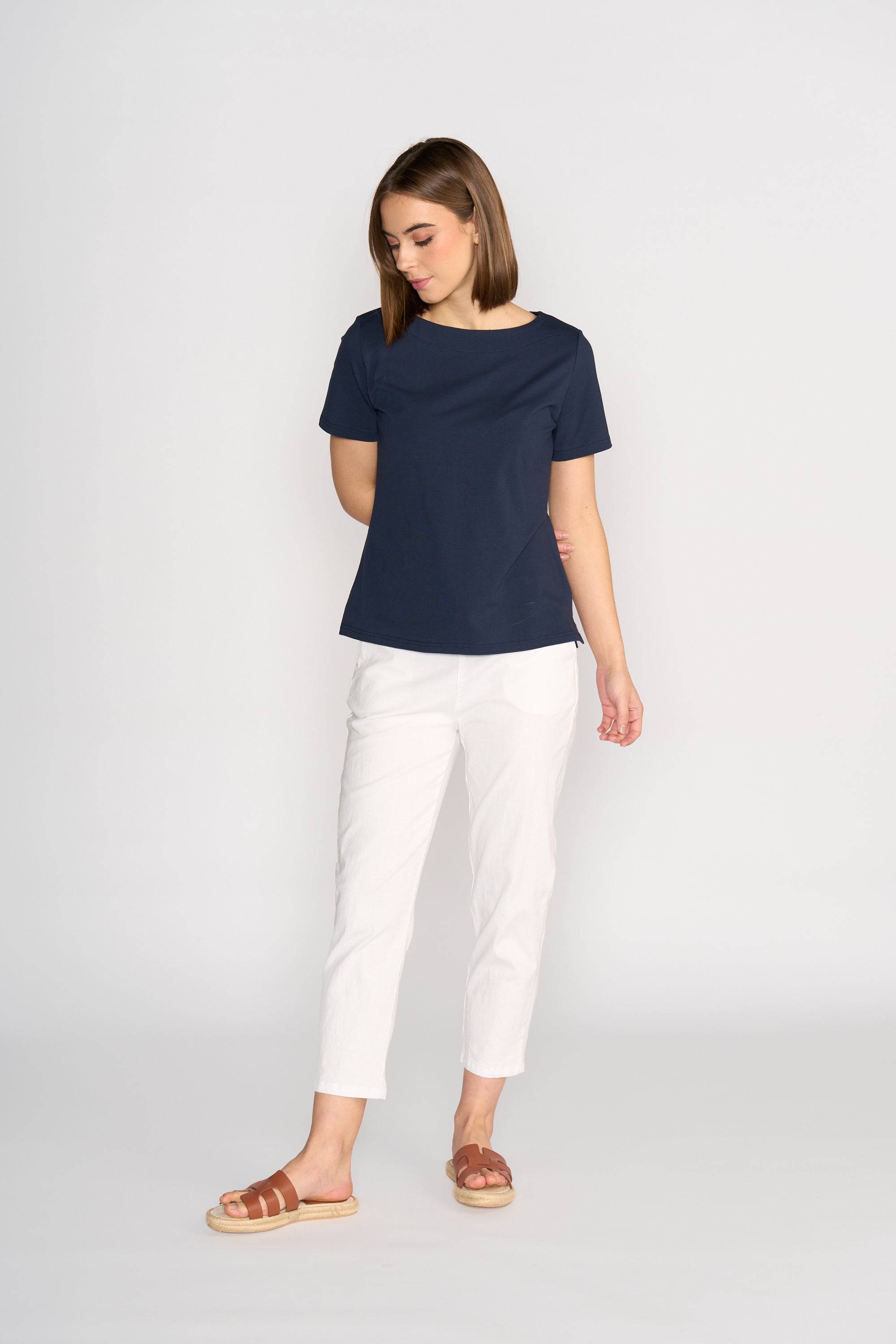A woman stands against a white background, wearing the Vassalli Raya Top in Ink. The top features a comfortable fit and boat neckline. She pairs it with white pants and brown sandals, looking down with her arms relaxed at her sides.