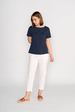 A woman stands against a white background, wearing the Vassalli Raya Top in Ink. The top features a comfortable fit and boat neckline. She pairs it with white pants and brown sandals, looking down with her arms relaxed at her sides.
