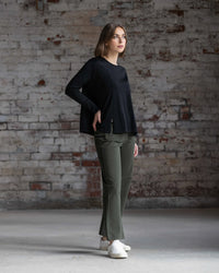 A woman stands indoors before a textured brick wall, wearing the Vassalli Pave Merino Top in Black, olive green pants, and white shoes. She looks to the side with one hand on her hip.