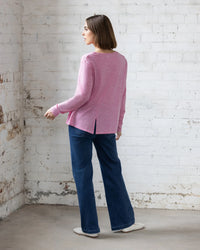 Indoors against a white brick wall, someone wears the Vassalli Pave Merino Top in Pink Marle, styled with blue wide-leg jeans and white shoes. The minimalist setting highlights this cozy, versatile piece—ideal for winter layering.