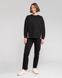 A woman with short brown hair smiles against a light gray background, wearing the Vassalli Zone Sweater in black, paired with black pants and light-colored sneakers.