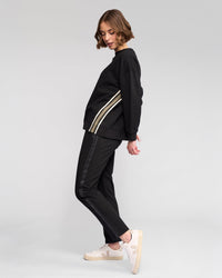 A woman poses sideways in a sporty Vassalli Zone Sweater - Black with white side stripes and matching black pants, paired with white sneakers against a plain light gray background.