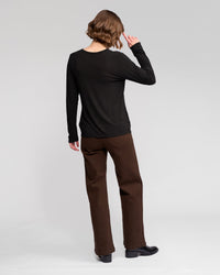 A person with short, wavy hair faces away, wearing the Vassalli Layer Top - Black paired with brown pants and black shoes against a plain light background.