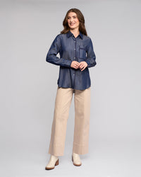 A woman wears the Vassalli Banner Shirt in deep blue, featuring embroidery, button front, and chest pocket, paired with wide-leg beige pants and cream ankle boots.