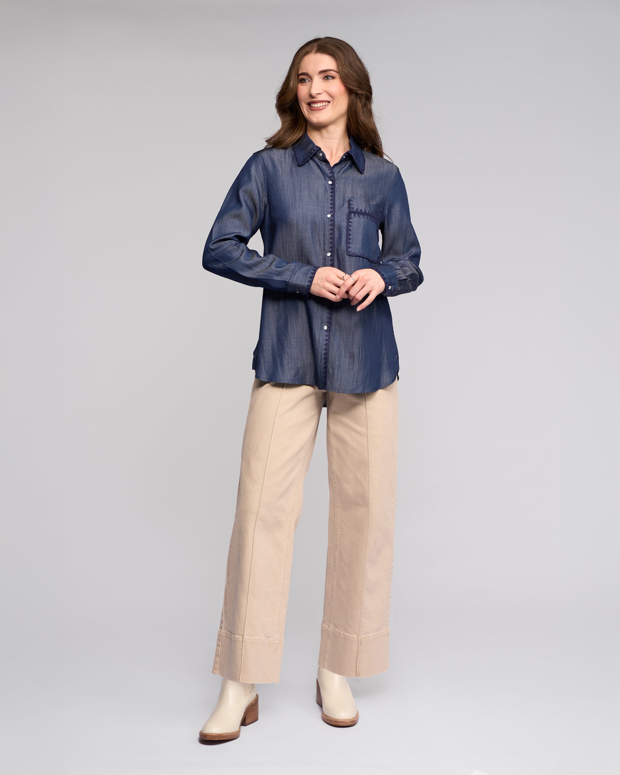 A woman wears the Vassalli Banner Shirt in deep blue, featuring embroidery, button front, and chest pocket, paired with wide-leg beige pants and cream ankle boots.