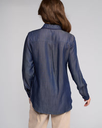 A person with long brown hair stands with their back to the camera, wearing the Vassalli Banner Shirt in Deep Blue and beige pants against a plain light gray background.