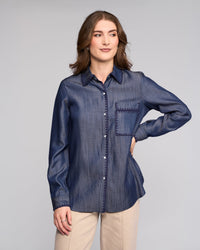 A woman with wavy brown hair models the Vassalli Banner Shirt in Deep Blue, featuring long sleeves and a front pocket, paired with beige pants. She stands with one hand on her hip against a plain light gray background.