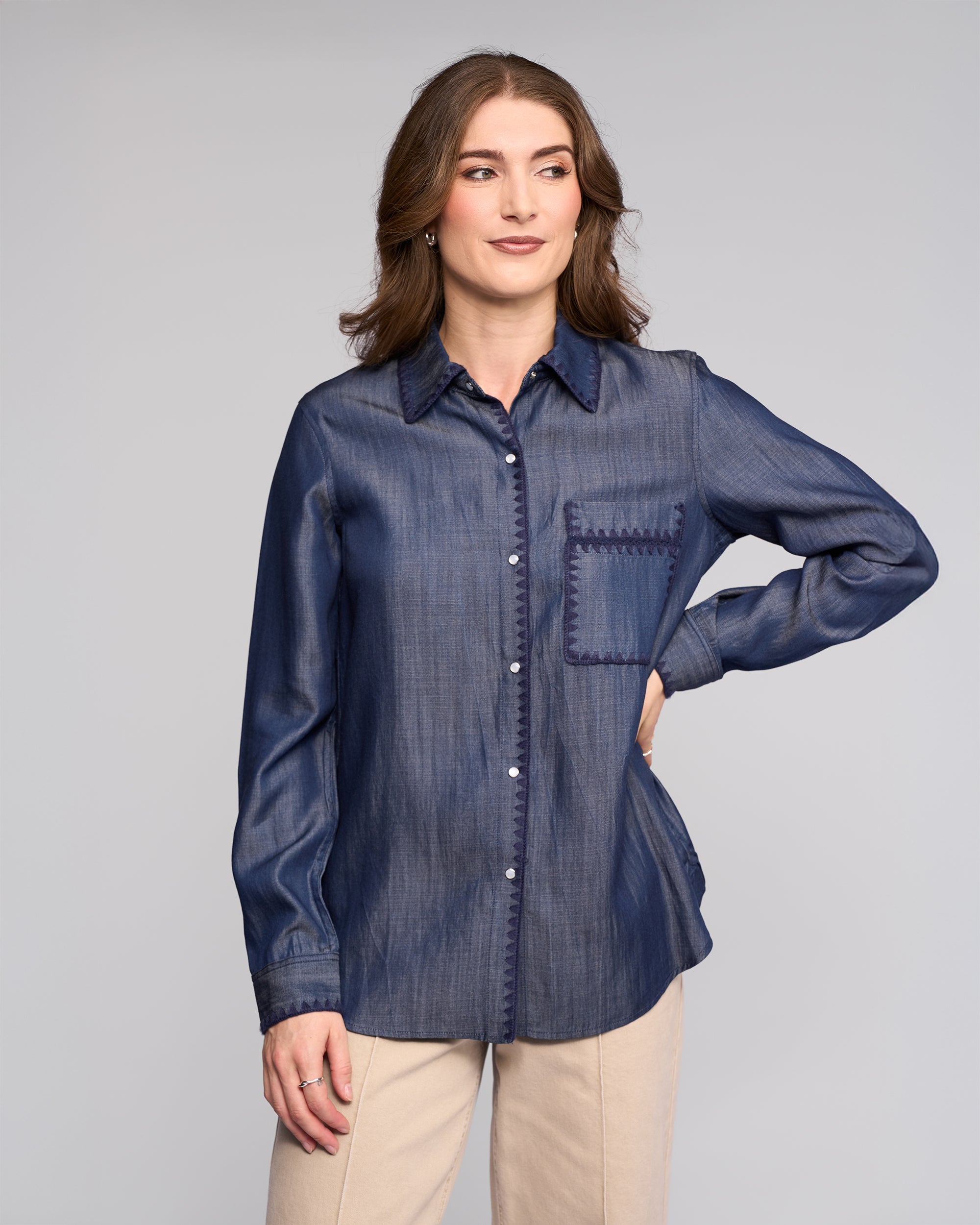 A woman with wavy brown hair models the Vassalli Banner Shirt in Deep Blue, featuring long sleeves and a front pocket, paired with beige pants. She stands with one hand on her hip against a plain light gray background.