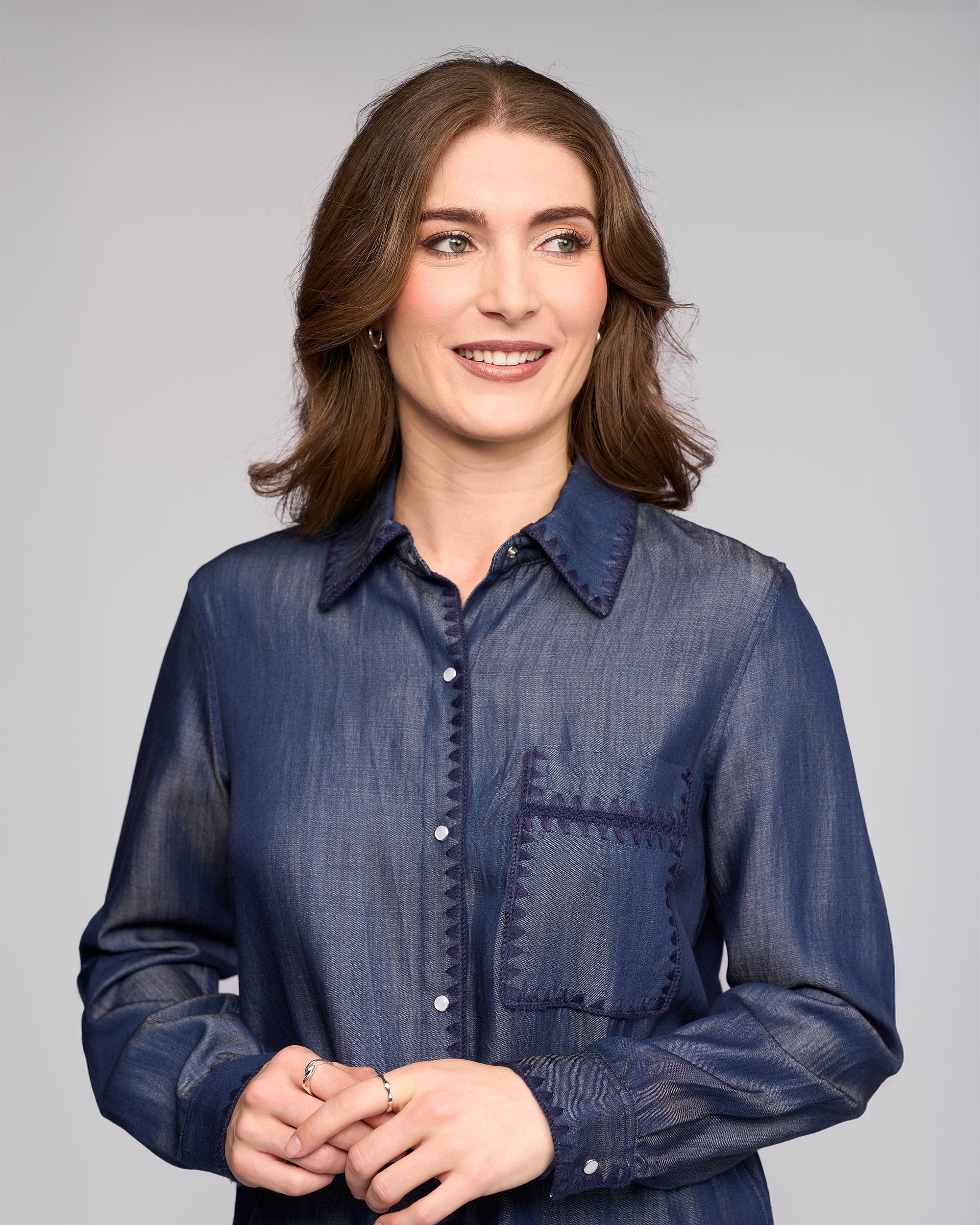 A woman with wavy brown hair, wearing the Vassalli Banner Shirt in deep blue, smiles and looks left with light makeup, hands together in front of her, set against a light gray background.