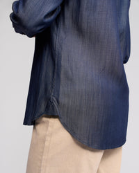 A person shown from the side at waist level against a plain background wears the Vassalli Banner Shirt in deep blue, featuring long sleeves, paired with light beige pants.