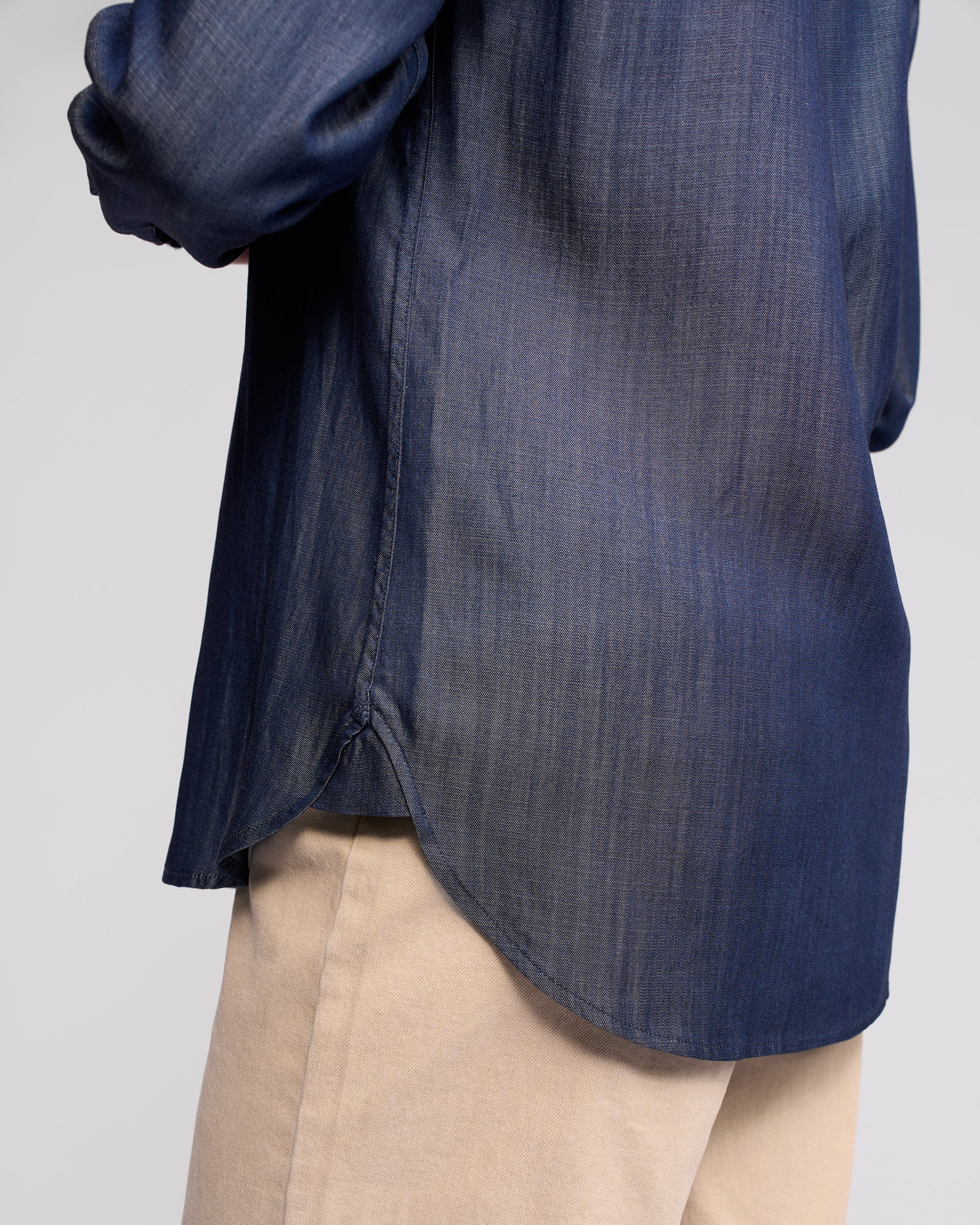 A person shown from the side at waist level against a plain background wears the Vassalli Banner Shirt in deep blue, featuring long sleeves, paired with light beige pants.