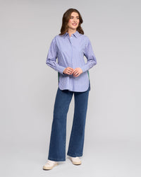 A woman smiles wearing the Vassalli Chart Shirt in Blue Stripe, an oversized cotton shirt, paired with blue wide-leg jeans and white sneakers against a plain light gray background.