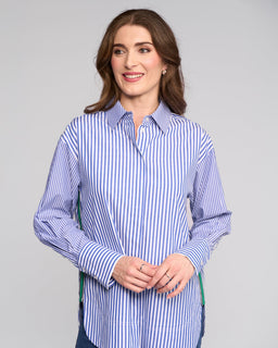 A woman with brown hair smiles gently in the Vassalli Chart Shirt - Blue Stripe, an oversized cotton piece, standing against a plain light gray background with her hands loosely clasped.
