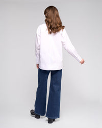 A person with wavy brown hair faces away, wearing the Vassalli Chart Shirt in white, paired with wide-leg blue jeans and black shoes, against a plain light background.