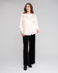 A woman with wavy brown hair and a neutral expression wears the Vassalli Charm Shirt in off white, featuring lace and black stitched details, paired with black flared pants and matching shoes against a plain background.