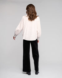 A person with wavy brown hair, seen from behind, wears the Vassalli Charm Shirt in off white with black stitched details and loose sleeves, paired with black wide-leg pants against a plain light background.