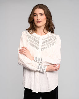 A woman with long brown hair models the Vassalli Charm Shirt in off white, featuring black stitched detailing. She stands against a light gray background, looking to the side with a relaxed expression and arms loosely crossed.
