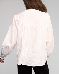 A person with long brown hair is seen from the back, wearing the Vassalli Charm Shirt in off white featuring black stitched cuffs, paired with black pants, against a plain light gray background.