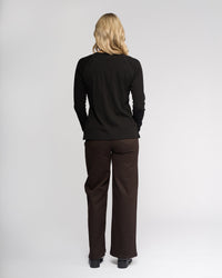 A person with long blonde hair, seen from behind, wears the Vassalli Key Top in black with dark brown pants and black shoes against a plain light background.