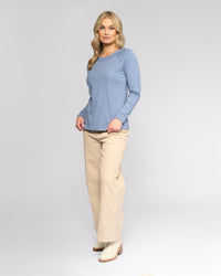 A woman with long blonde hair stands against a light background, wearing the Vassalli Key Top in denim blue, beige wide-leg pants, and cream ankle boots. She faces forward with a relaxed expression.