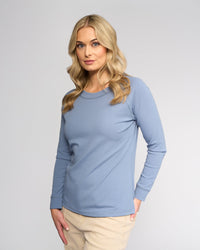 A woman with long blonde hair wears the Vassalli Key Top in denim blue, paired with beige pants, standing against a plain light gray background and looking slightly to the side with a neutral expression.