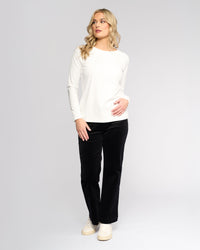 A woman with long blonde hair wears the Vassalli Key Top in Winter White, styled casually with black pants and white sneakers, standing against a light background and looking left with one hand on her hip.