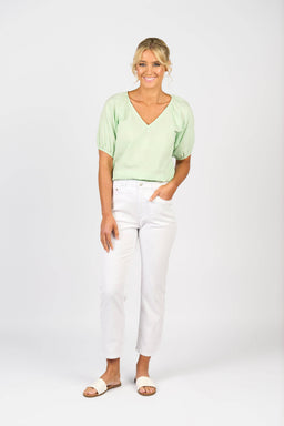 A woman stands against a white background, smiling. She wears a pale green blouse, Vassalli Onya Jean - White Denim ankle grazers with star embroidery, and white sandals. One hand is in her pocket; her blond hair is styled up.