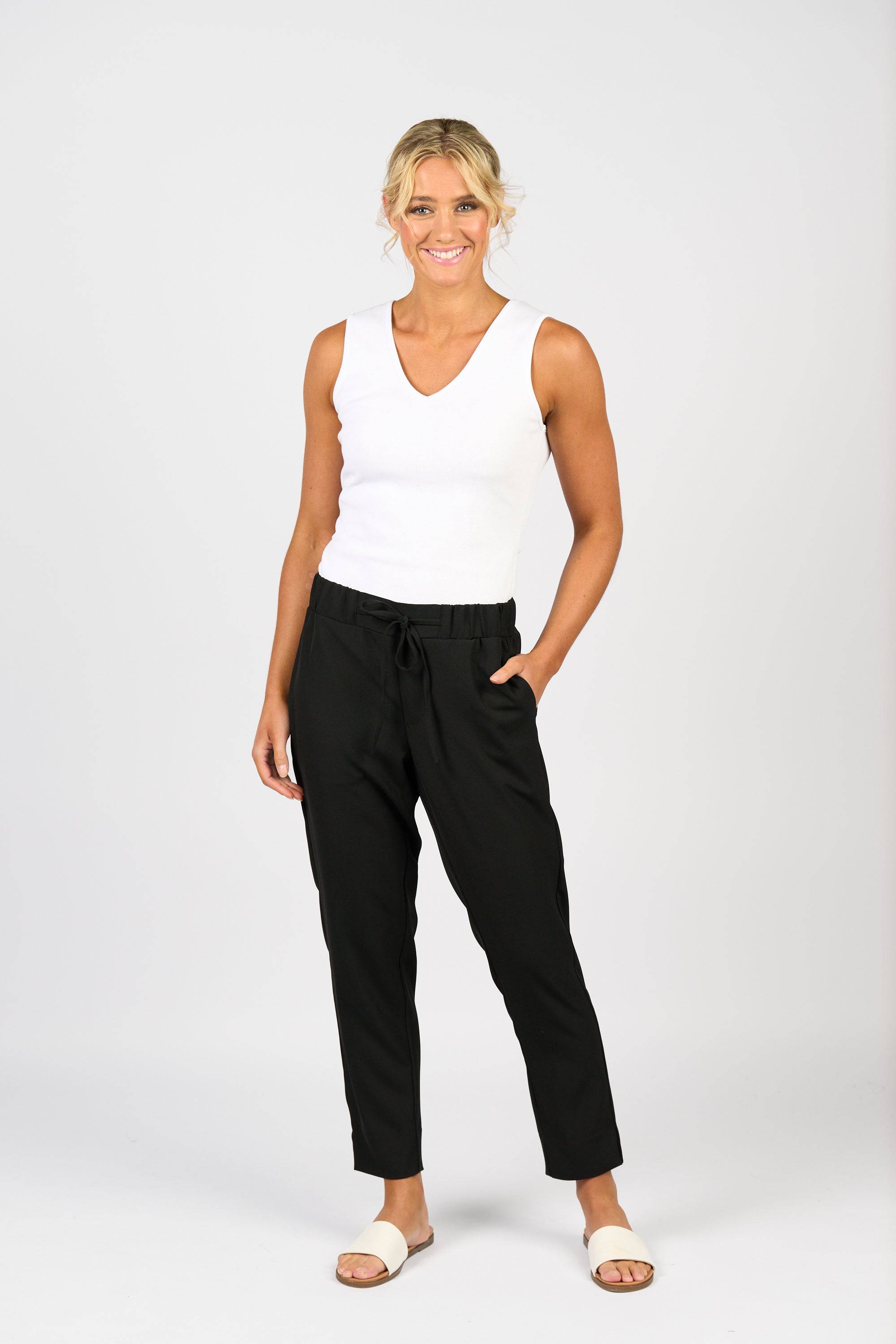 A woman smiles against a white background, wearing Vassalli’s Marble Pant in Black with an elastic waistband and a white sleeveless top. One hand is in her pocket, the other by her side, paired with white sandals.