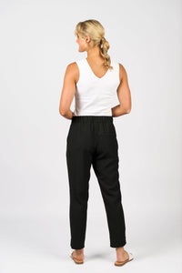 A woman with a blonde ponytail faces away, wearing a white sleeveless top, Vassalli Marble Pant - Black with an elastic waistband, and beige sandals against a plain white background.