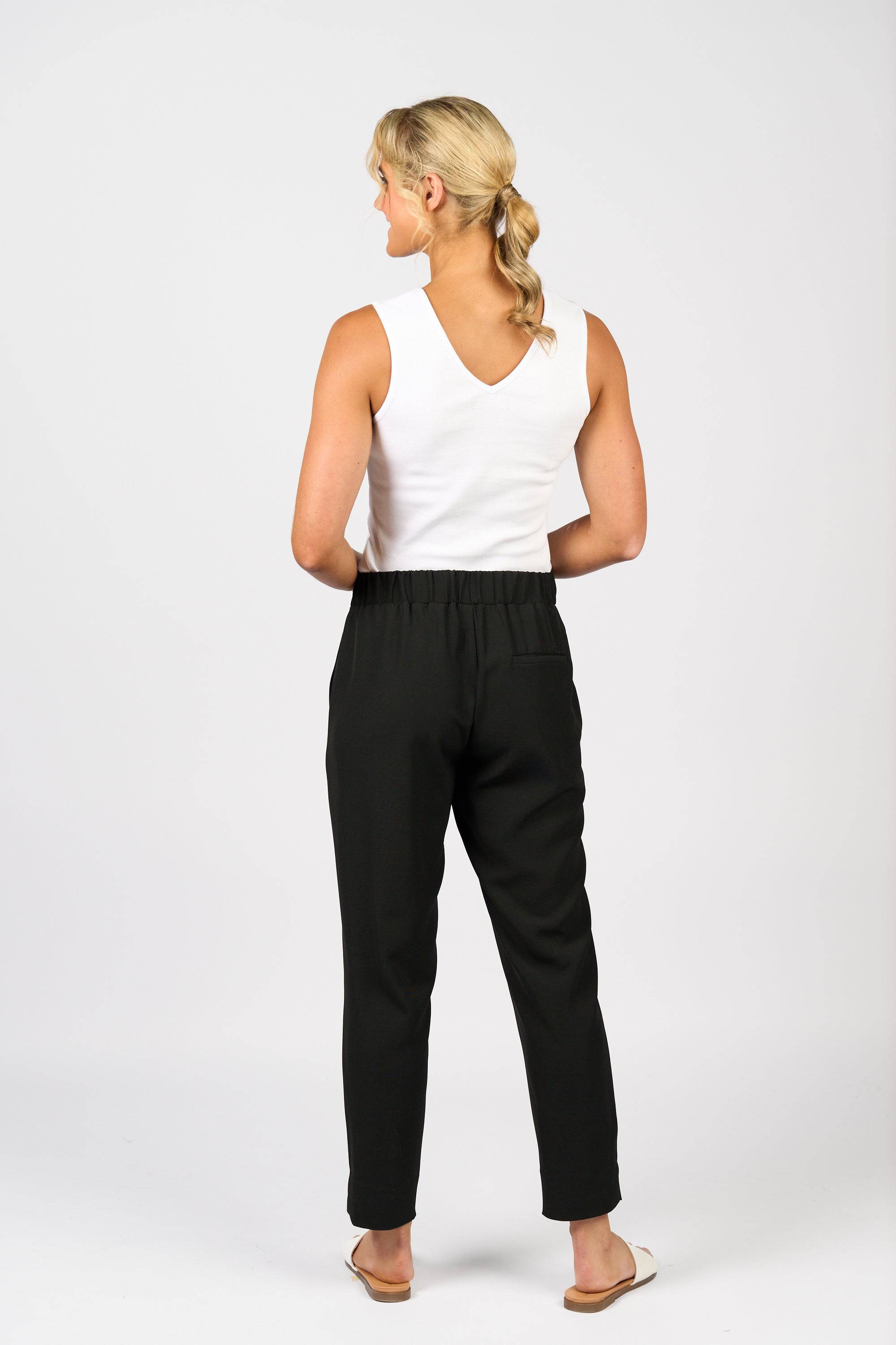 A woman with a blonde ponytail faces away, wearing a white sleeveless top, Vassalli Marble Pant - Black with an elastic waistband, and beige sandals against a plain white background.