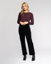 A woman with wavy blonde hair wears the Vassalli Market Cord Pant in black, paired with a maroon long-sleeve top and black boots. She stands against a plain light gray background, smiling slightly at the camera.