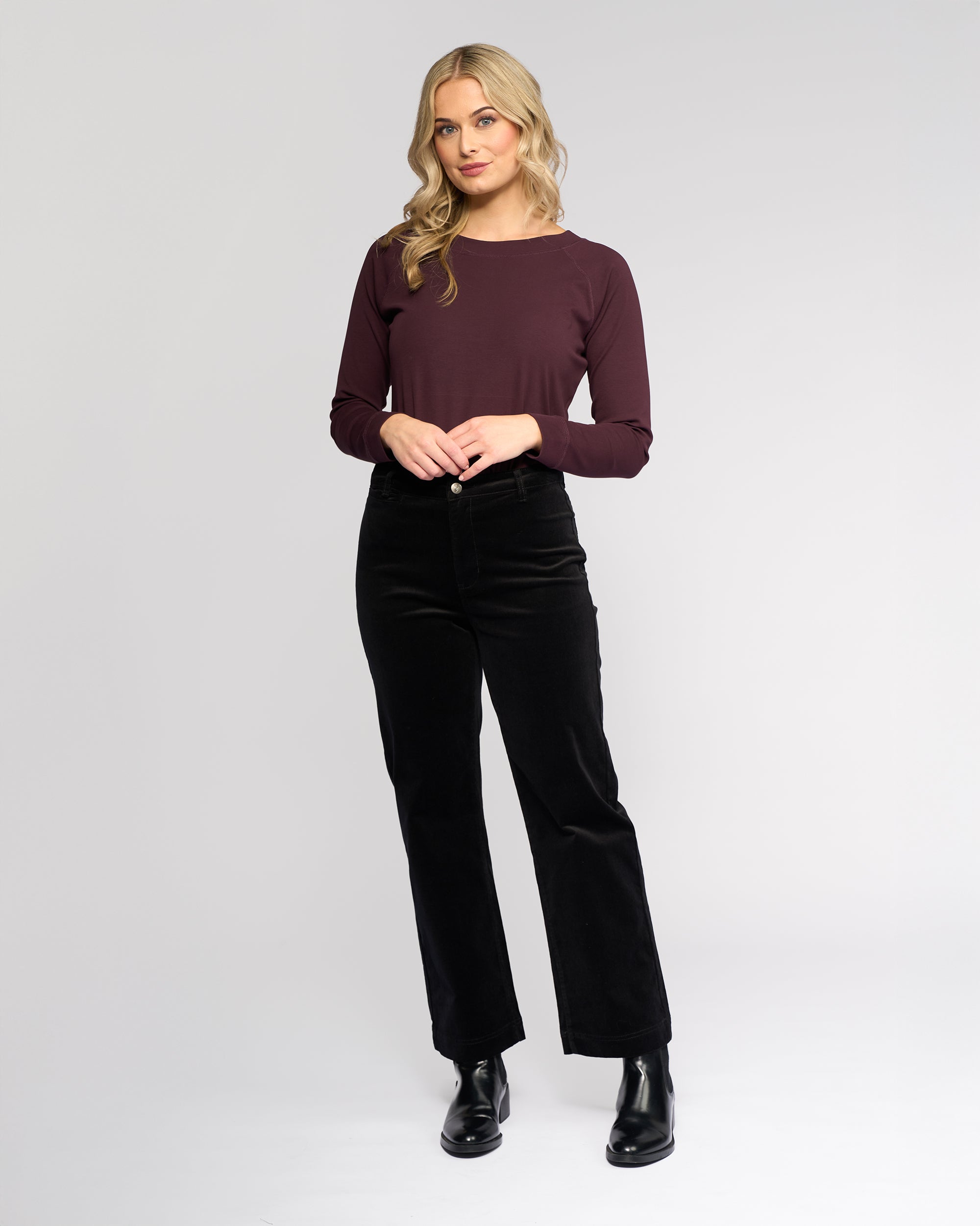 A woman with wavy blonde hair wears the Vassalli Market Cord Pant in black, paired with a maroon long-sleeve top and black boots. She stands against a plain light gray background, smiling slightly at the camera.