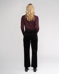 A woman with long blonde hair, seen from behind, wears a maroon long-sleeve top and Vassalli Market Cord Pant in black, paired with black shoes, standing against a plain light gray background.