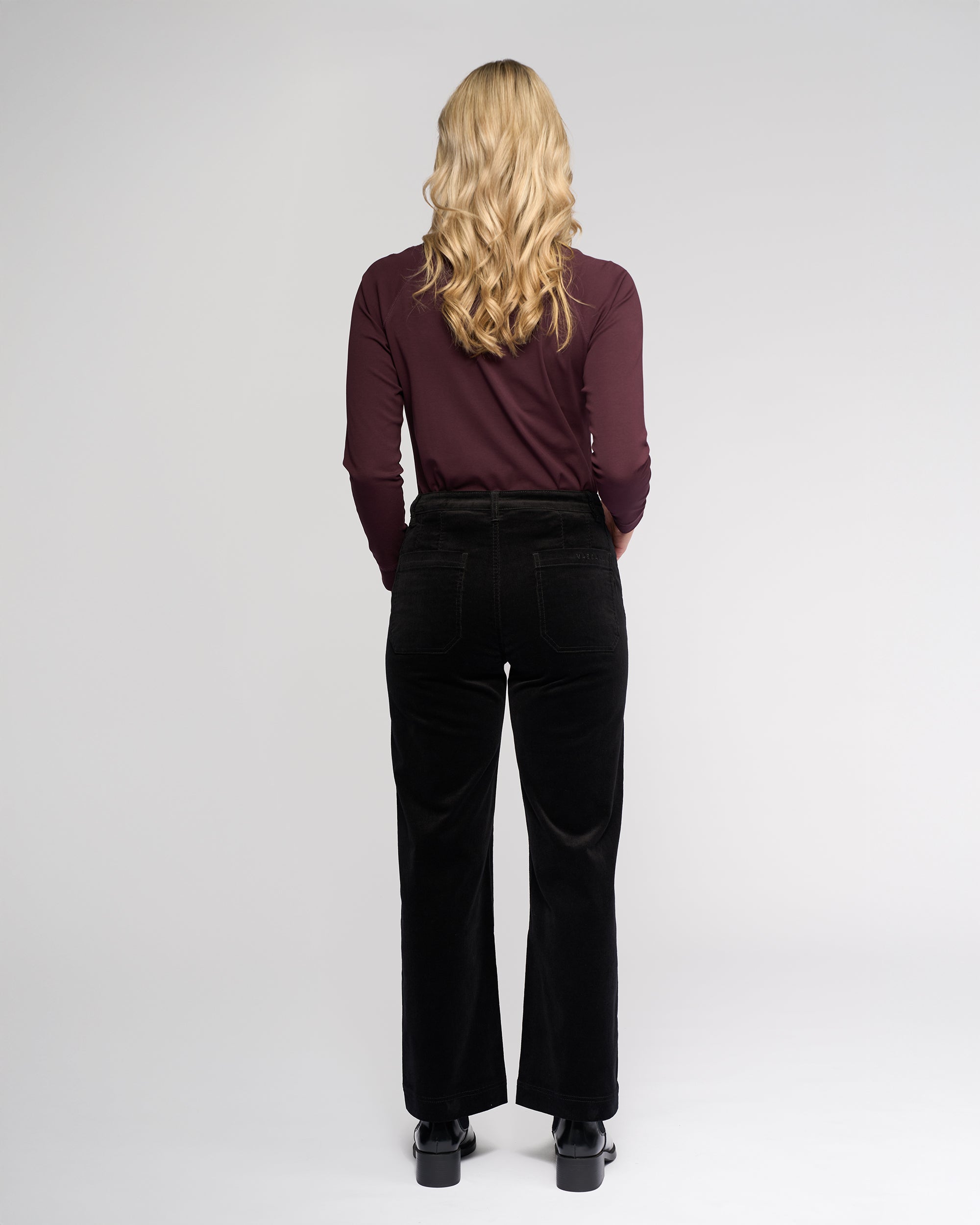 A woman with long blonde hair, seen from behind, wears a maroon long-sleeve top and Vassalli Market Cord Pant in black, paired with black shoes, standing against a plain light gray background.