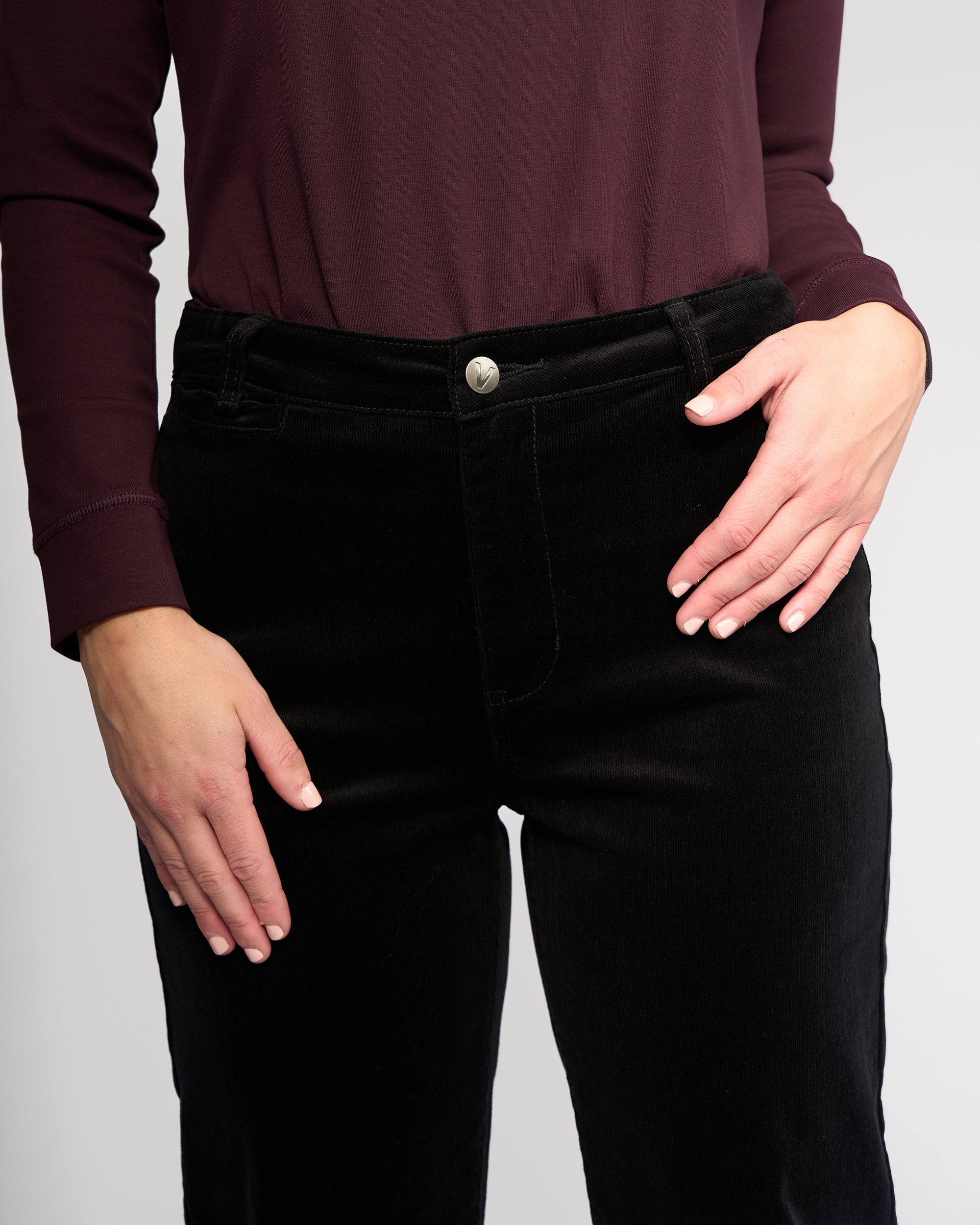 A person models the Vassalli Market Cord Pant in Black, featuring a relaxed-leg ankle-grazer fit. They're also wearing a long-sleeved maroon top. The image is cropped from shoulders to thighs, with one hand on their hip and the other by their side.