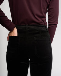 Person wearing Vassalli's Market Cord Pant in black, shown from the back with one hand on hip. Pants feature stretch fabric, visible back pockets, and a subtle brand logo above the right pocket.