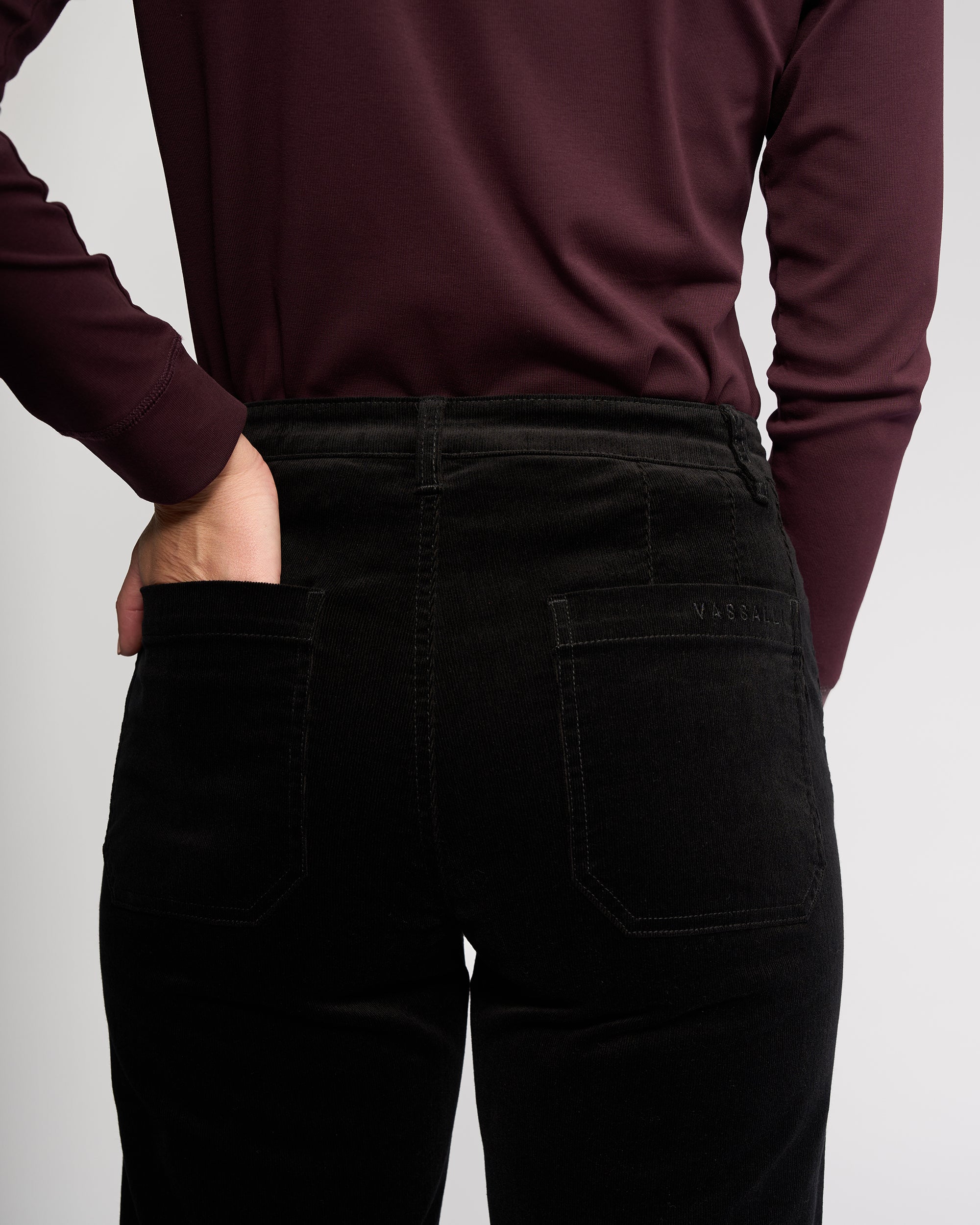 Person wearing Vassalli's Market Cord Pant in black, shown from the back with one hand on hip. Pants feature stretch fabric, visible back pockets, and a subtle brand logo above the right pocket.