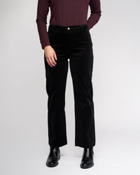 A person stands against a plain background wearing a long-sleeve burgundy top, Vassalli Market Cord Pant in black, and black ankle boots. Their hands are in their pockets, and their head is out of frame.