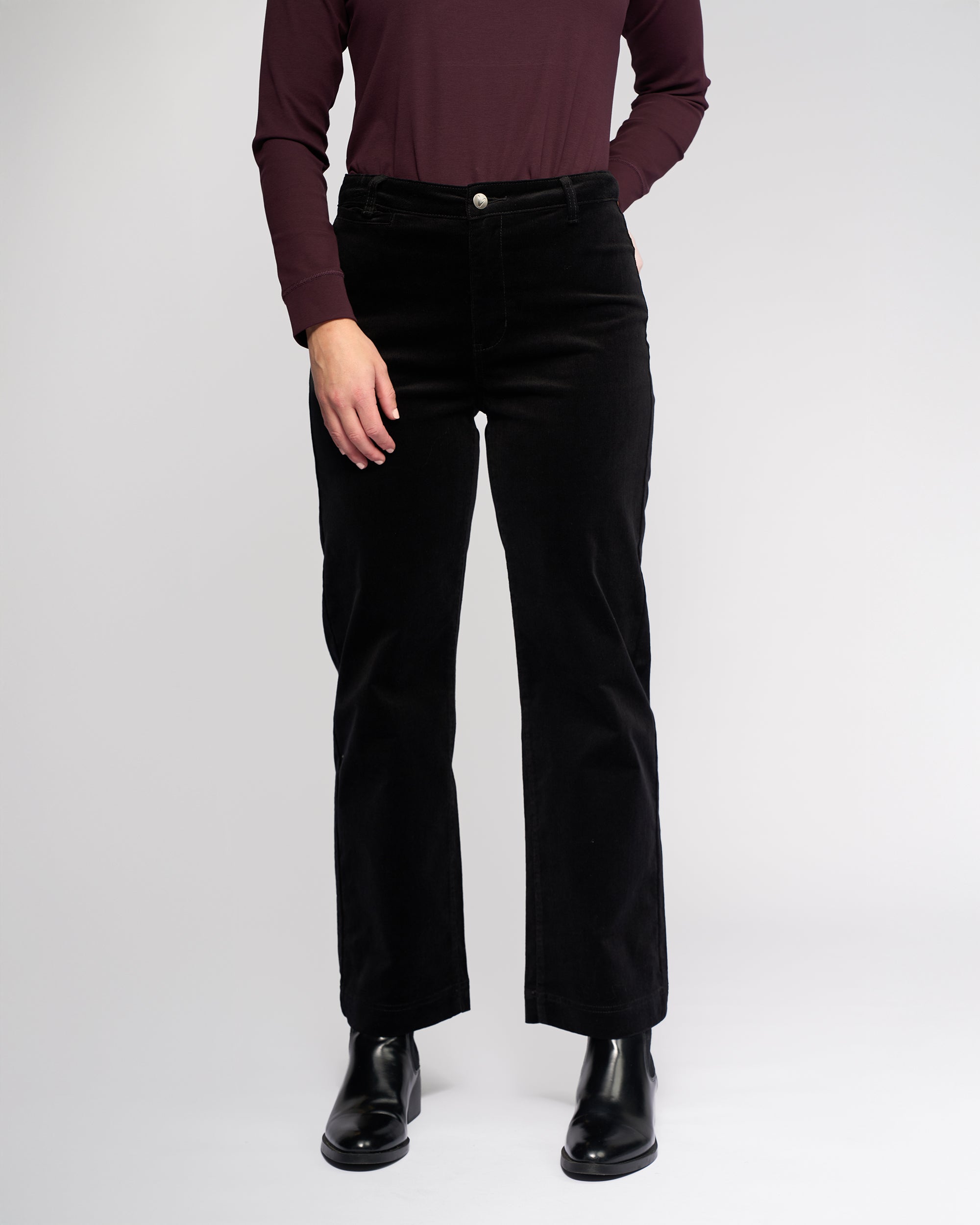 A person stands against a plain background wearing a long-sleeve burgundy top, Vassalli Market Cord Pant in black, and black ankle boots. Their hands are in their pockets, and their head is out of frame.