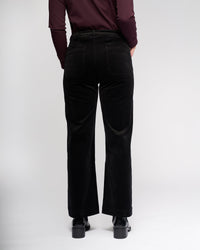 A person stands facing away from the camera, wearing Vassalli Market Cord Pant in black with a long-sleeve burgundy top and black shoes, set against a plain white background.