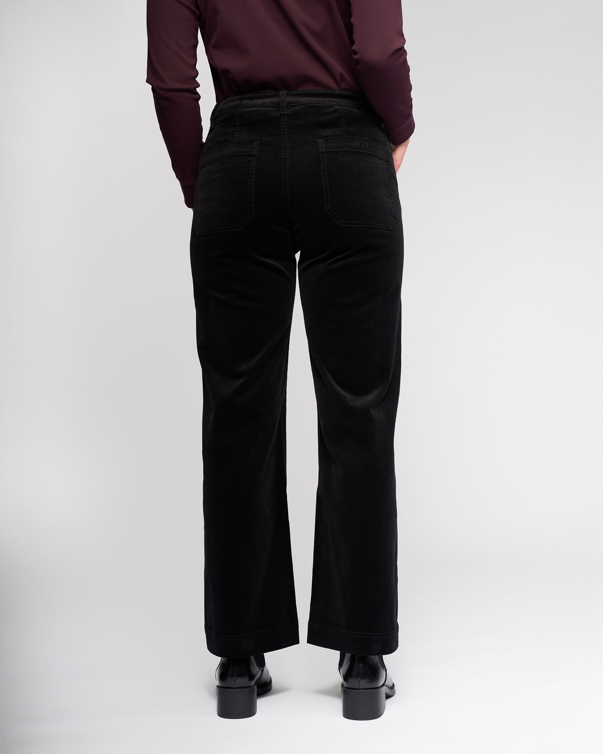 A person stands facing away from the camera, wearing Vassalli Market Cord Pant in black with a long-sleeve burgundy top and black shoes, set against a plain white background.