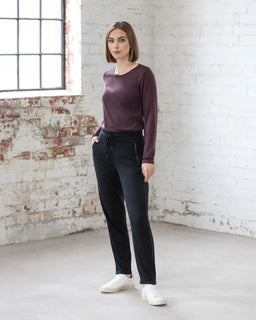 A woman stands indoors by a large window, wearing the Vassalli Walker Merino Pant in Black, paired with a maroon long-sleeve top and white sneakers. The backdrop is a white-painted brick wall with exposed brick patches.