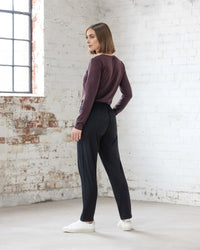 A woman with chin-length brown hair stands in a bright, industrial-style room with white brick walls. She wears a maroon top, Vassalli Walker Merino Pant in black, and white sneakers, looking over her shoulder at the camera.