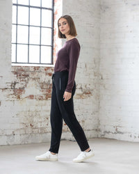 A woman with a bob haircut, wearing a maroon long-sleeve top, white sneakers, and Vassalli's Walker Merino Pant in black, stands sideways in a bright room with white brick walls and large windows.