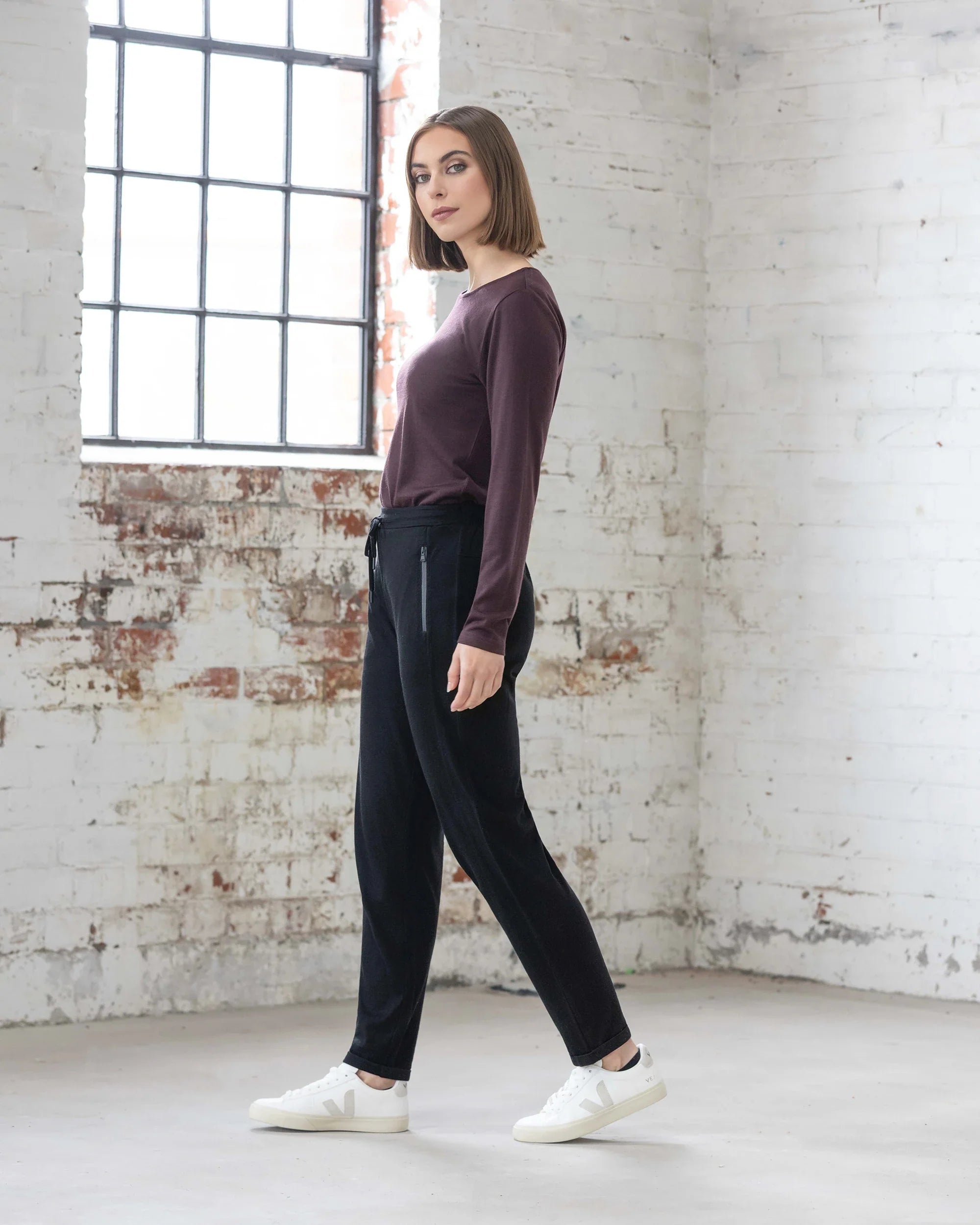 A woman with a bob haircut, wearing a maroon long-sleeve top, white sneakers, and Vassalli's Walker Merino Pant in black, stands sideways in a bright room with white brick walls and large windows.