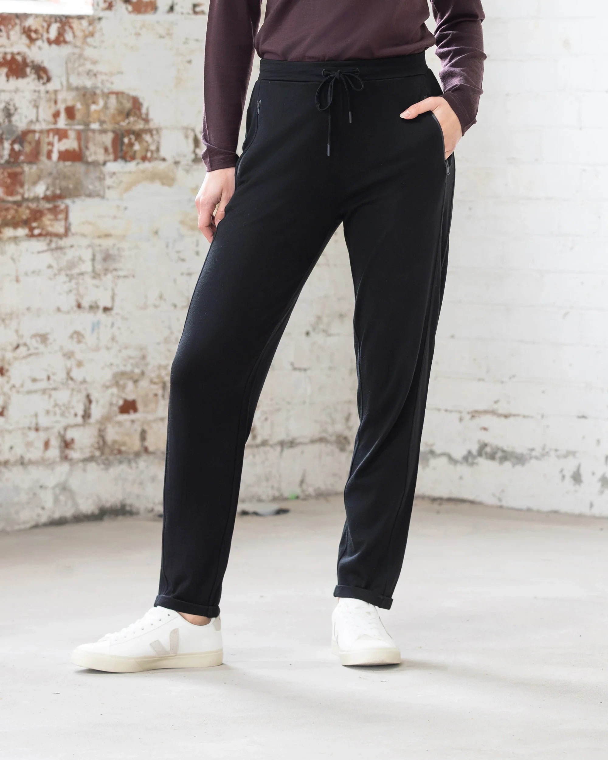 A person stands indoors against a white brick wall, wearing Vassalli Walker Merino Pant in black, a long-sleeve maroon top, and white sneakers. One hand is in their pocket; only their body from the shoulders down is visible.